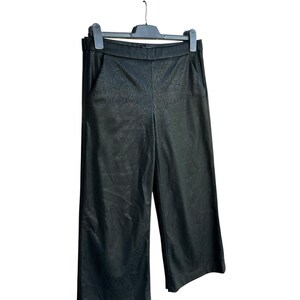 Puede incluir: Pantalones culotte negros de piel sintética con una sutil textura de piel de serpiente, colgados de una percha negra. Los pantalones tienen un diseño de pierna ancha y dos bolsillos delanteros. El material parece tener un acabado mate.