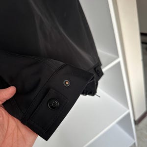 K&ouml;nnte beinhalten: Nahaufnahme eines schwarzen Jacken&auml;rmels. Der &Auml;rmel hat eine Manschette mit einem schwarzen Knopf und einem kleineren, kreisf&ouml;rmigen Knopf. Der Stoff wirkt glatt und matt. Im Hintergrund ist ein wei&szlig;es Regal zu sehen. Die Jacke ist teilweise sichtbar.