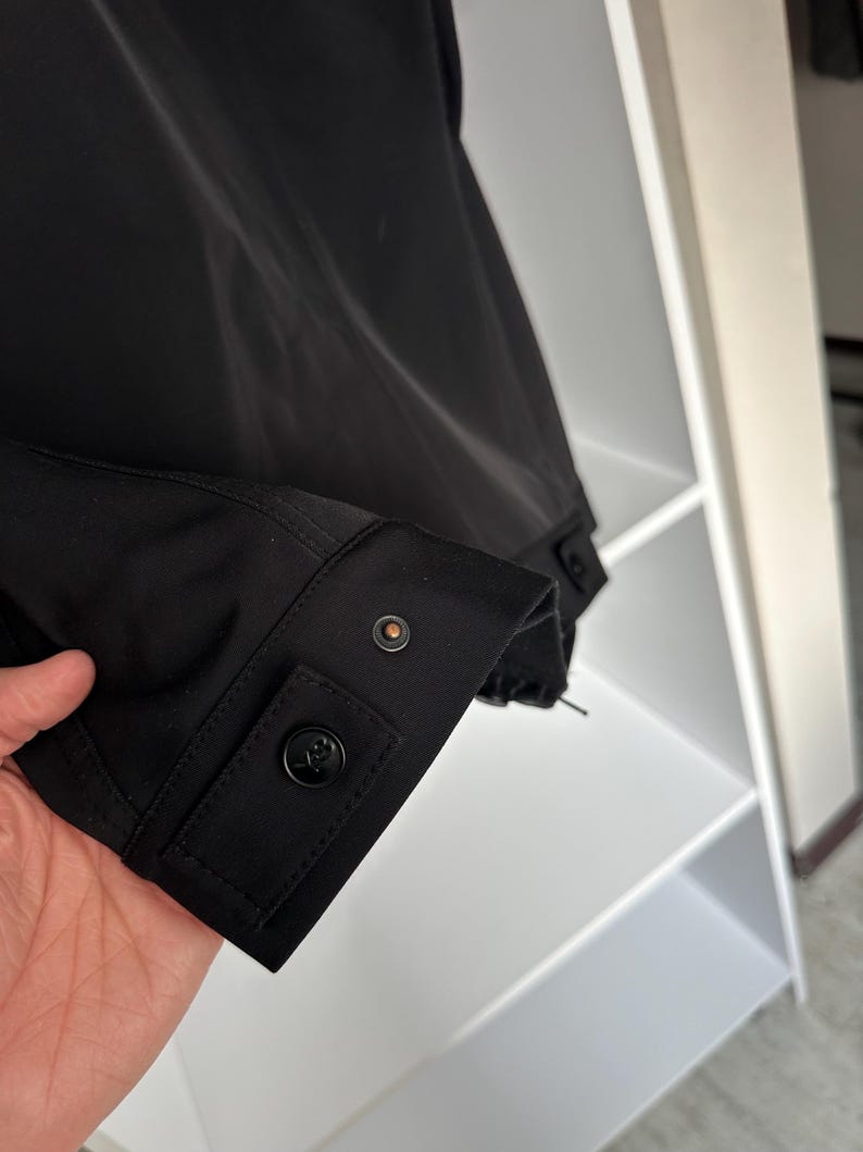 K&ouml;nnte beinhalten: Nahaufnahme eines schwarzen Jacken&auml;rmels. Der &Auml;rmel hat eine Manschette mit einem schwarzen Knopf und einem kleineren, kreisf&ouml;rmigen Knopf. Der Stoff wirkt glatt und matt. Im Hintergrund ist ein wei&szlig;es Regal zu sehen. Die Jacke ist teilweise sichtbar.