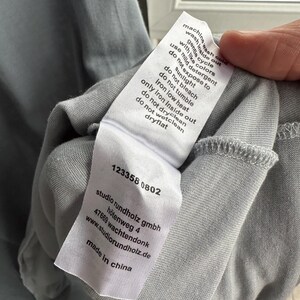 以下が含まれることがあります： ライトブルーの衣服のクローズアップ。白いタグには洗濯表示と「studio rundholz gmbh」と「made in china」の文字が記載されています。