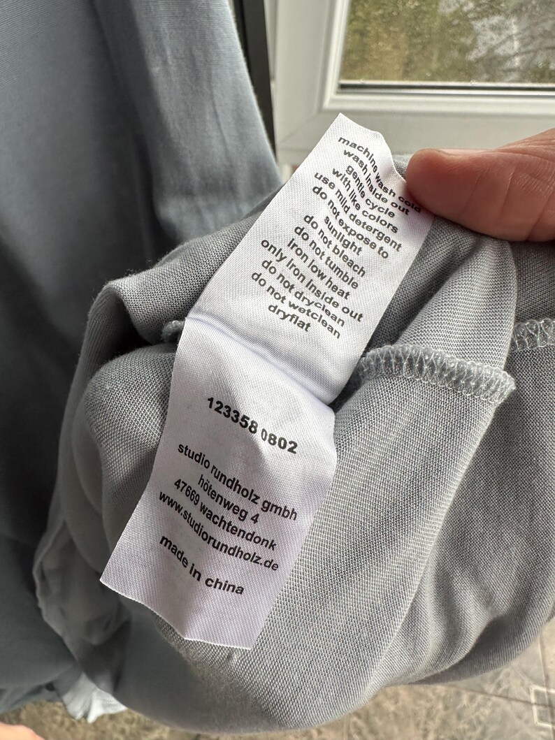 以下が含まれることがあります： ライトブルーの衣服のクローズアップ。白いタグには洗濯表示と「studio rundholz gmbh」と「made in china」の文字が記載されています。