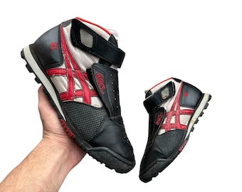 May include: Black, red, and white high-top trainers with a velcro strap. The trainers feature the brand name "Asics" and a red stripe design on the side. The soles are black and white.