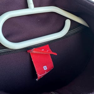 May include: A dark purple athletic jacket with a black zip and a red tag with a white "S" on it. The jacket is hanging on a white plastic hanger.