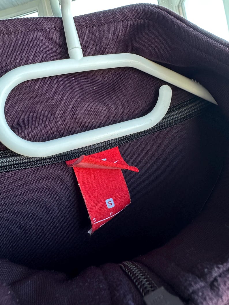 May include: A dark purple athletic jacket with a black zip and a red tag with a white "S" on it. The jacket is hanging on a white plastic hanger.