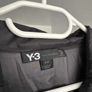 K&ouml;nnte beinhalten: Schwarze Y-3 Jacke an einem wei&szlig;en Kleiderb&uuml;gel. Die Jacke hat ein schwarzes Etikett mit wei&szlig;em Text: "Y-3 YOHJI YAMAMOTO adidas". Das Etikett gibt auch die Gr&ouml;&szlig;e S/P und "MADE IN CHINA" an.