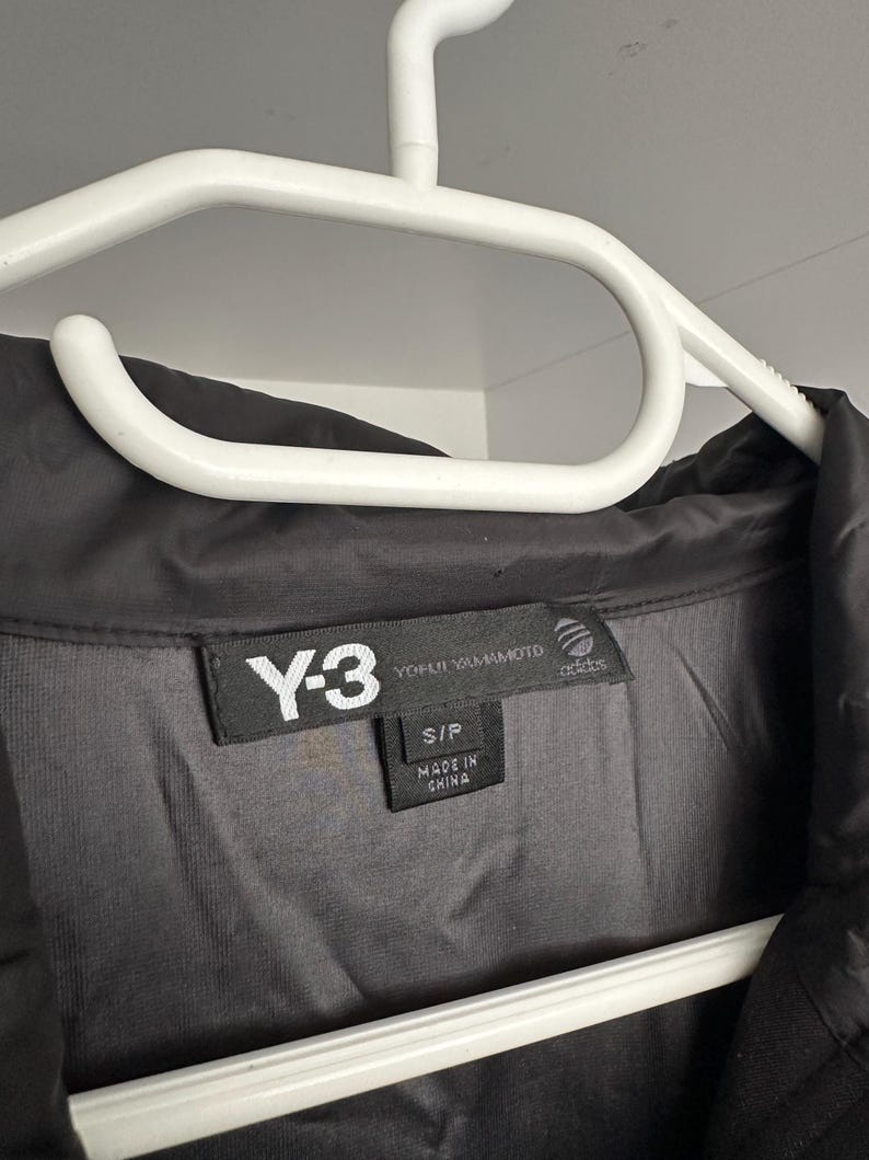 K&ouml;nnte beinhalten: Schwarze Y-3 Jacke an einem wei&szlig;en Kleiderb&uuml;gel. Die Jacke hat ein schwarzes Etikett mit wei&szlig;em Text: "Y-3 YOHJI YAMAMOTO adidas". Das Etikett gibt auch die Gr&ouml;&szlig;e S/P und "MADE IN CHINA" an.