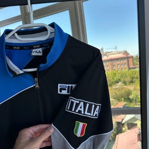May include: A black, blue, and gray zip-up jacket with the word "ITALIA" and an Italian flag patch on the sleeve. The jacket has the "FILA" logo on the chest and collar.