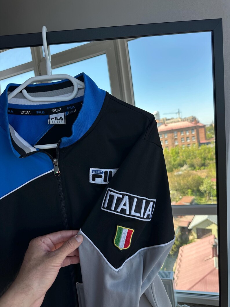 May include: A black, blue, and gray zip-up jacket with the word "ITALIA" and an Italian flag patch on the sleeve. The jacket has the "FILA" logo on the chest and collar.
