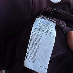 May include: A dark purple athletic jacket with a zipper and a Galatasaray emblem. The jacket has a white tag with text in multiple languages, including "100% POLYESTER". The emblem features the number "1905".