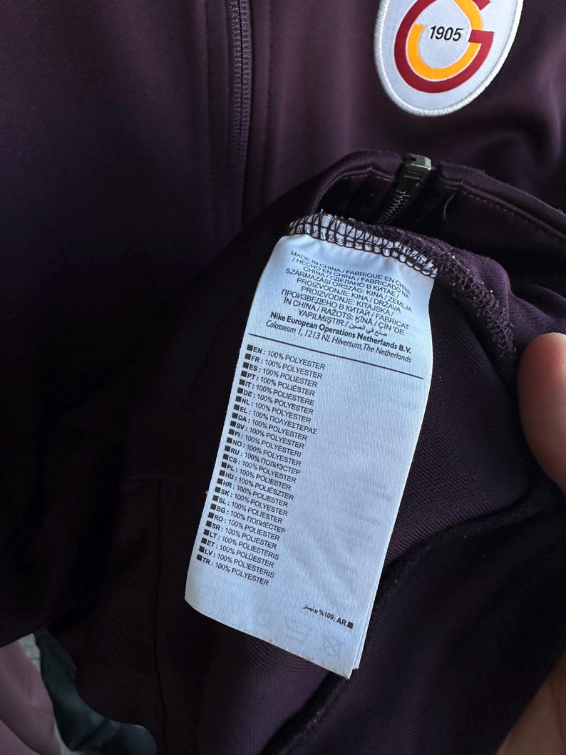 May include: A dark purple athletic jacket with a zipper and a Galatasaray emblem. The jacket has a white tag with text in multiple languages, including "100% POLYESTER". The emblem features the number "1905".