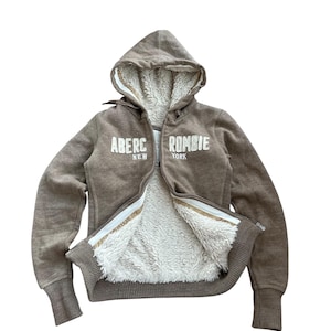 May include: A brown zip-up hoodie with a white faux-fur lining. The front features the text ABERCROMBIE NEW YORK in white embroidered lettering. The garment includes a hood with drawstrings, long sleeves with ribbed cuffs, and a ribbed hem.