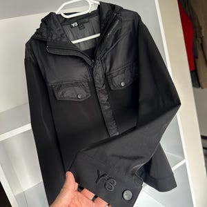 K&ouml;nnte beinhalten: Schwarze Y-3 Jacke mit Kapuze, zwei Brusttaschen und gekn&ouml;pften Manschetten. Das Y-3-Logo ist auf der Manschette gestickt. Die Jacke h&auml;ngt an einem wei&szlig;en Kleiderb&uuml;gel in einem wei&szlig;en Schrank.