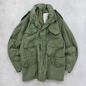 Può includere: Giacca verde oliva in stile militare con multiple tasche frontali e chiusura con bottoni. La giacca ha un colletto e spalline sulle spalle. Il tessuto sembra essere un materiale resistente, possibilmente cotone.