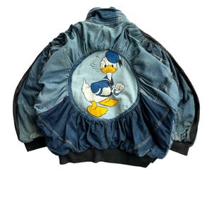 Może przedstawiać: Kurtka dżinsowa z dużą okrągłą naszywką przedstawiającą Kaczora Donalda w stroju marynarza. Kurtka ma niebieski dżinsowy korpus z czarnymi rękawami i czarnym prążkowanym dołem. Tył kurtki ma falbaniasty dżinsowy detal wokół naszywki Kaczora Donalda.