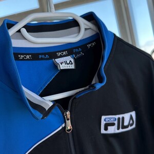 May include: A black and blue Fila sport jacket with a white zipper and collar. The jacket has the word "SPORT" printed on the collar and the Fila logo on the chest. The jacket is hanging on a white hanger.