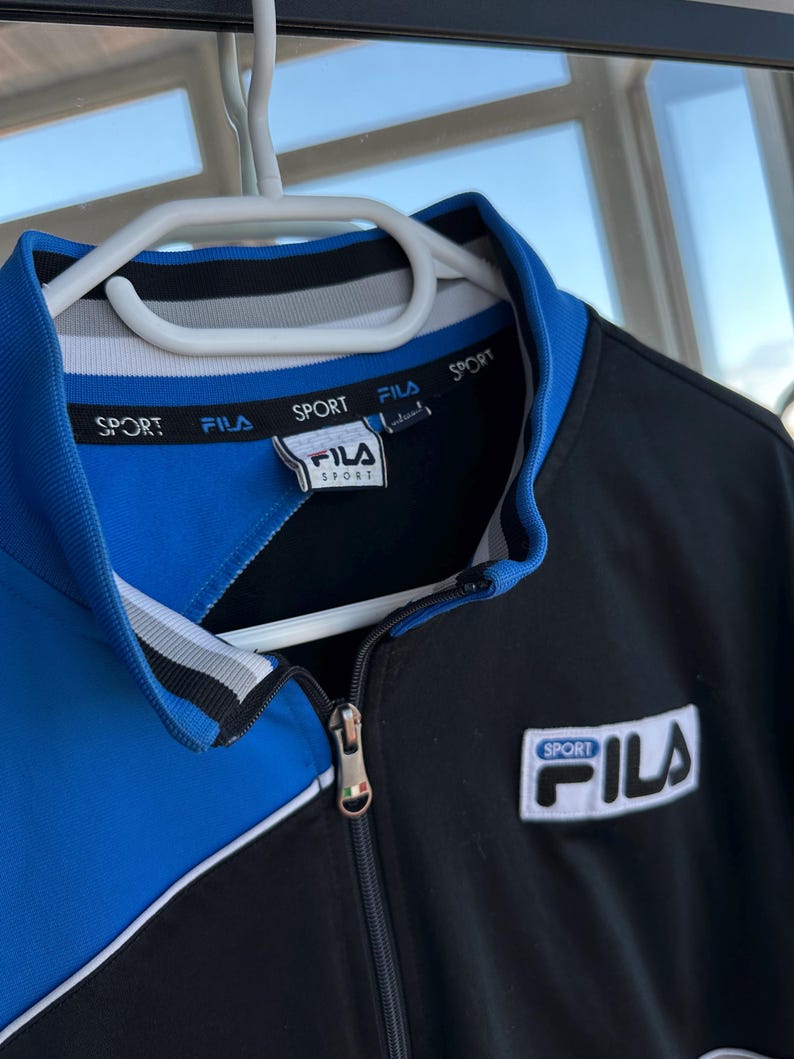 May include: A black and blue Fila sport jacket with a white zipper and collar. The jacket has the word "SPORT" printed on the collar and the Fila logo on the chest. The jacket is hanging on a white hanger.