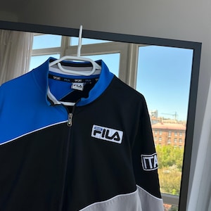 May include: A Fila track jacket in black, blue, and gray, hanging on a white hanger. The jacket has a full zipper, a stand-up collar, and the Fila logo on the chest. The sleeve has an "ITA" patch.