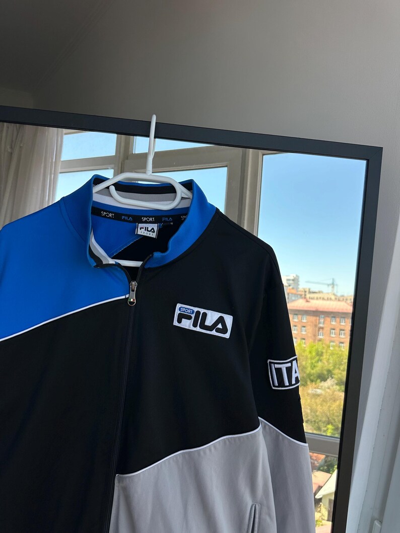 May include: A Fila track jacket in black, blue, and gray, hanging on a white hanger. The jacket has a full zipper, a stand-up collar, and the Fila logo on the chest. The sleeve has an "ITA" patch.