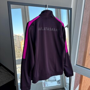 May include: A dark purple Galatasaray track jacket with a high collar and a full-length zip. The jacket has bright pink stripes down the sleeves and the word "GALATASARAY" printed across the back in a lighter shade of purple.