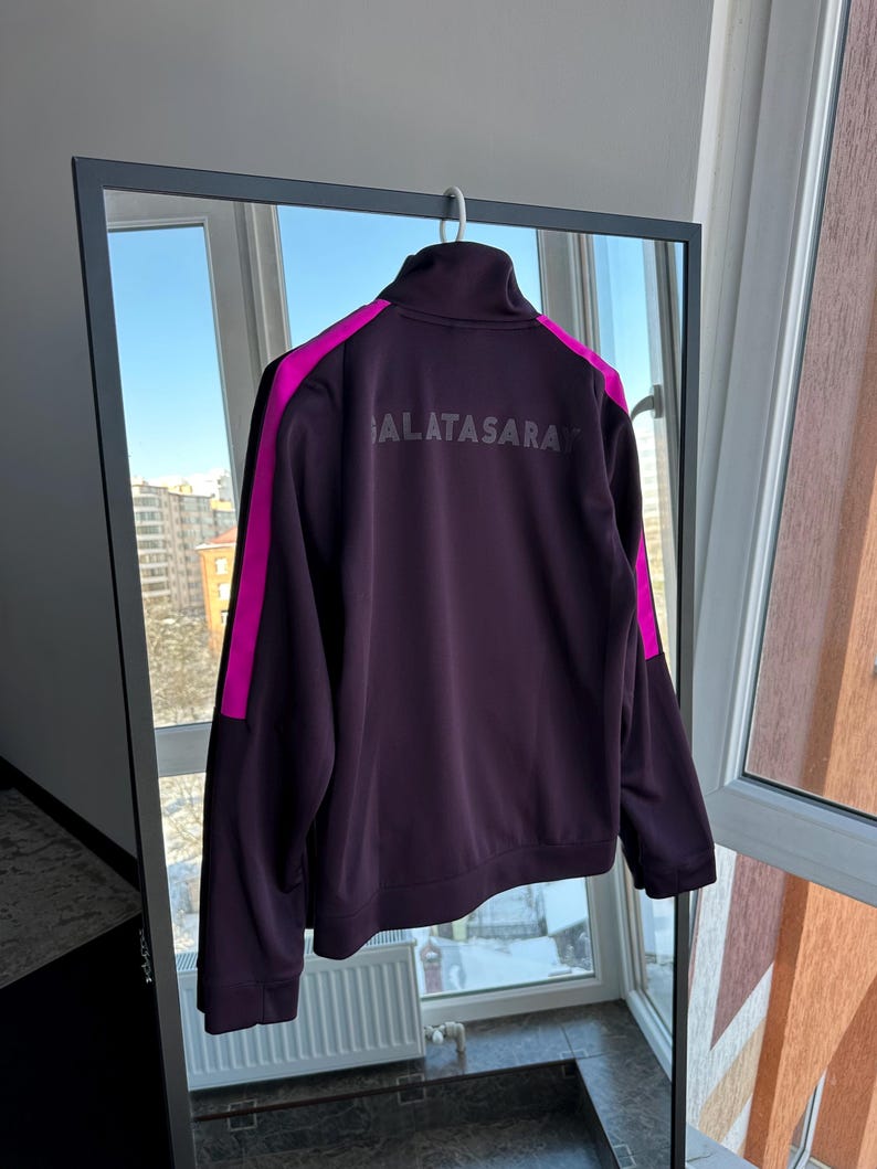 May include: A dark purple Galatasaray track jacket with a high collar and a full-length zip. The jacket has bright pink stripes down the sleeves and the word "GALATASARAY" printed across the back in a lighter shade of purple.
