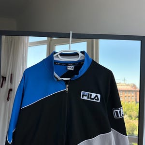 May include: A Fila track jacket in black, blue, and gray, hanging on a white hanger. The jacket features a full zipper, a blue collar, and the Fila logo on the chest. The Italian flag is on the sleeve.