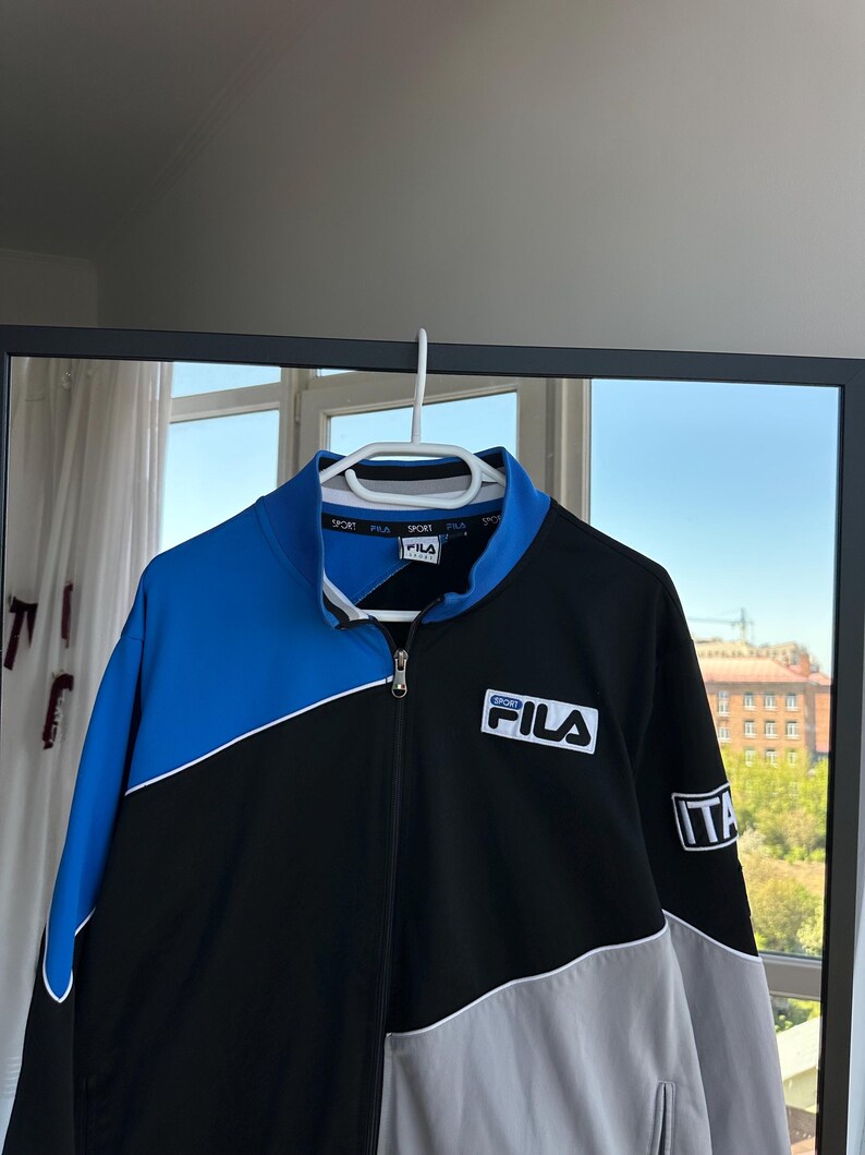 May include: A Fila track jacket in black, blue, and gray, hanging on a white hanger. The jacket features a full zipper, a blue collar, and the Fila logo on the chest. The Italian flag is on the sleeve.