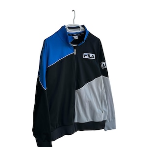May include: A Fila track jacket in black, blue, and gray, hanging on a white hanger. The jacket features a full zipper, a high collar, and the Fila logo on the chest. The word "ITA" is on the sleeve.