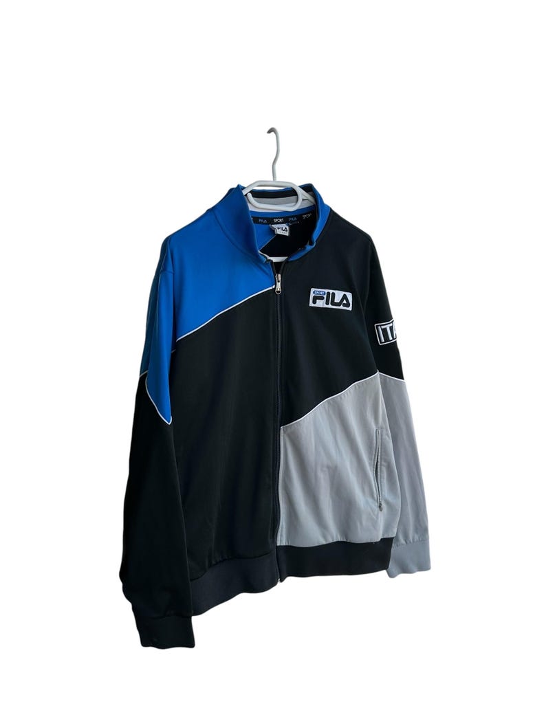 May include: A Fila track jacket in black, blue, and gray, hanging on a white hanger. The jacket features a full zipper, a high collar, and the Fila logo on the chest. The word "ITA" is on the sleeve.