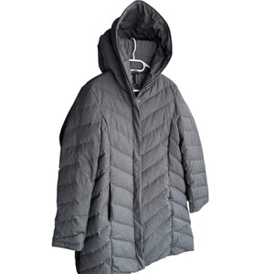 May include: A gray hooded winter coat with a chevron quilted pattern. The coat has a zipper closure and two pockets. The hood is attached and the coat is hanging on a white hanger.