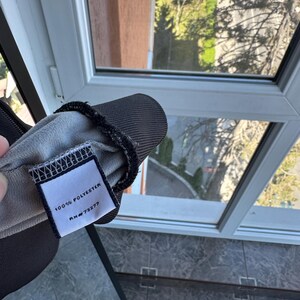 May include: Close-up of a dark gray fabric item with a tag. The tag reads "100% POLYESTER" and "RN #73277". The item has a black trim and a light gray interior. The background shows a window with a view of trees and a building.