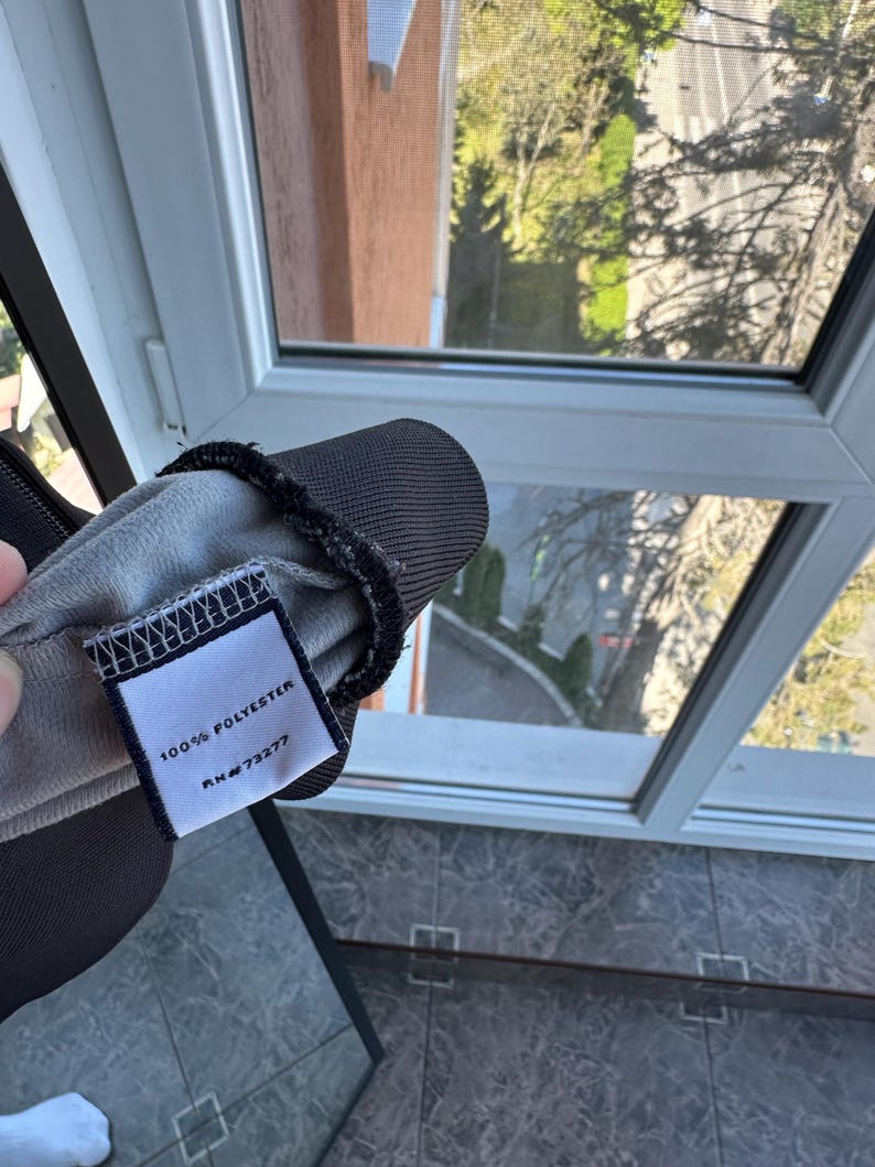 May include: Close-up of a dark gray fabric item with a tag. The tag reads "100% POLYESTER" and "RN #73277". The item has a black trim and a light gray interior. The background shows a window with a view of trees and a building.