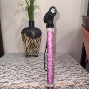 Könnte beinhalten: Dekorative Gehhilfe mit schwarzem Griff und einem rosa, strassbesetzten Schaft. Der Gehstock hat ein kleines Licht am Griff und eine Handschlaufe. Der Schaft ist mit rosa Strasssteinen bedeckt.