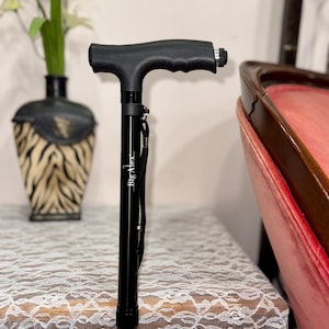 May include: Black adjustable walking cane with a textured handle and the text "Big Alex". The bottom of the cane has a colorful beaded design. The cane is resting on a white lace tablecloth.