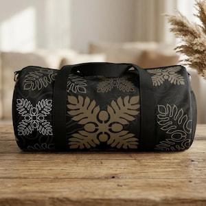 May include: A black duffel bag featuring a repeating pattern of tan and white stylized fern leaf motifs. The bag includes black fabric handles and a central strap, with a cylindrical shape and a smooth, durable exterior finish.