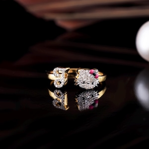 May include: A gold ring with a Hello Kitty design featuring a white face with black eyes and a pink bow. The ring is set with clear cubic zirconia stones.