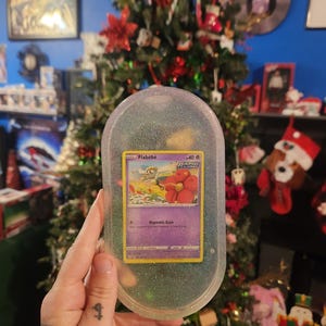 May include: A clear, glittery resin case holding a Flabebe Pokémon card. The card features a cartoon flower and creature with the text "Hypnotic Gaze". The case is oval-shaped and has a Christmas tree in the background.