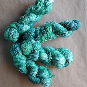 May include: A collection of hand-dyed yarn skeins in teal, turquoise, and white hues. The yarn is arranged in a flowing pattern against a neutral brown backdrop. The yarn appears soft and suitable for knitting or crochet projects.