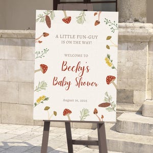 May include: A baby shower welcome sign on a wooden easel. The sign features the text "A LITTLE FUN-GUY IS ON THE WAY!" and "WELCOME TO Becky's Baby Shower" with the date August 16, 2025. The sign is decorated with mushroom and leaf illustrations.