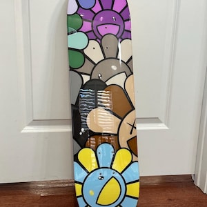 May include: A colorful skateboard featuring a repeating pattern of smiling flower faces in various colors. The flowers have yellow, blue, purple, and brown petals. The skateboard is a standard size and shape.