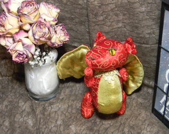 Puede incluir: Un dragón de peluche rojo y dorado con alas. El dragón tiene un diseño detallado y un solo ojo amarillo.