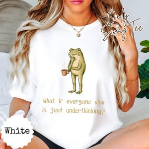 May include: A white t-shirt featuring a graphic illustration of a green frog holding a steaming brown mug. Below the frog, the text reads, "What if everyone else is just underthinking?" The shirt has a crew neckline and short sleeves.