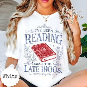 May include: A white t-shirt featuring a graphic print with the text I'VE BEEN READING SINCE THE LATE 1900s. The design includes a red book illustration centered between the text, surrounded by blue decorative flourishes and leaf patterns.