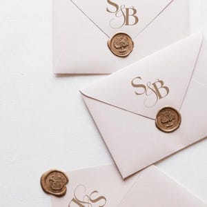 Könnte beinhalten: Drei rosafarbene Umschläge mit goldenen Siegeln aus Wachs und den aufgedruckten Initialen "S&B". Die Wachssiegel zeigen ein Baummotiv.