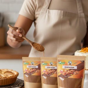Puede incluir: Una persona esparce especias para pastel de una cuchara de madera sobre tres bolsas. Las bolsas están etiquetadas como "CAKE SPICE" y presentan imágenes de productos horneados. También se ve un pastel y una tarta.