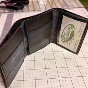 May include: A black tri-fold wallet constructed from recycled rubber material. The interior features multiple card slots and a clear plastic ID window containing a business card with the text Rubber Revolution, Pike Place Market, Miranda Marks, Creator.