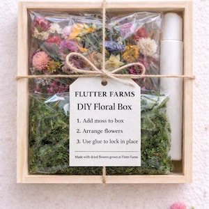 May include: A square wooden box containing a clear plastic bag filled with colorful dried flowers and green moss, alongside a white glue bottle. A tag tied with twine reads: FLUTTER FARMS DIY Floral Box, 1. Add moss to box, 2. Arrange flowers, 3. Use glue to lock in place. Made with dried flowers grown at Fluter Farms.
