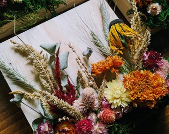 Könnte beinhalten: Eine quadratische Holzplatte, verziert mit einem Arrangement aus Trockenblumen, darunter gelbe, orangefarbene und rosafarbene Blüten, grünes Moos, Eukalyptusblätter und Weizenhalme. Das Blumenarrangement ist auf der hellen Holzoberfläche befestigt.