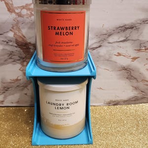 May include: Two scented candles in glass jars, one labeled "Strawberry Melon" and the other "Laundry Room Lemon", sit on a blue tiered stand. The Strawberry Melon candle has a red label with white text. The Laundry Room Lemon candle has a white label.