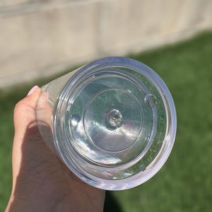 Pode incluir: Um copo de pl&aacute;stico transparente com um fundo liso e arredondado. O copo &eacute; segurado na m&atilde;o.