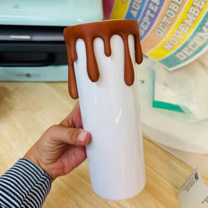 May include: A white cylindrical tumbler featuring a brown drip design around the top rim. The brown material appears to be a textured, raised coating that mimics liquid dripping down the sides of the smooth, white surface of the drink container.